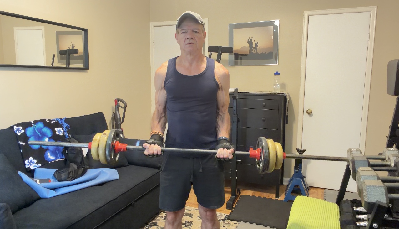 John standing tall with a barbell at the lowered starting position of a biceps curl