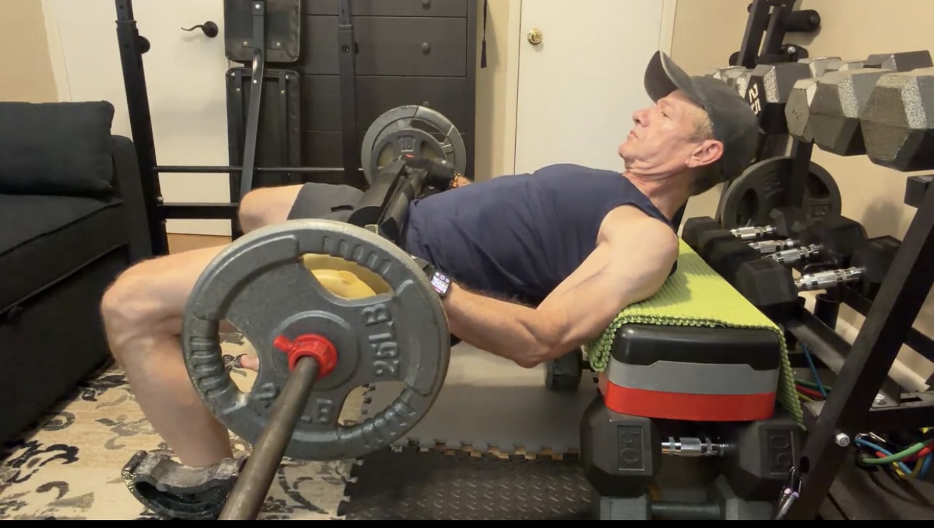 John performing a barbell hip thrust in his home gym at age 64